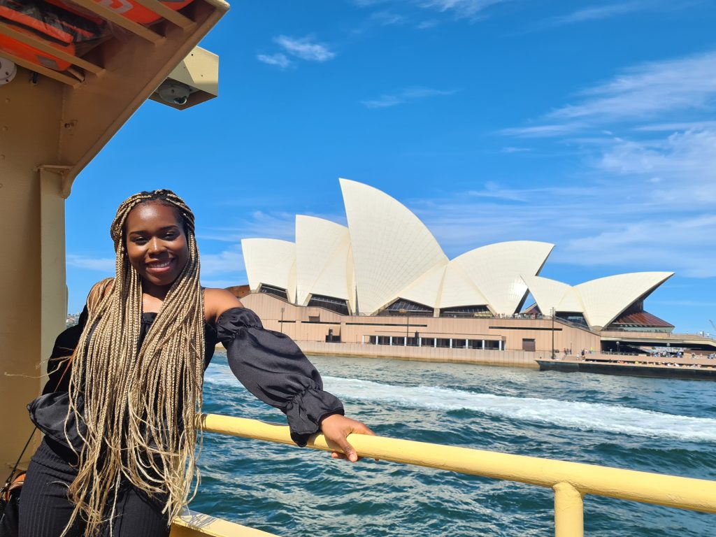 Young woman posing on a boat with the Sydney Opera House behind her.