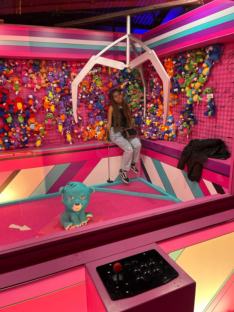 Young woman in a fake claw machine setting with the claw over her head and toys in the background.